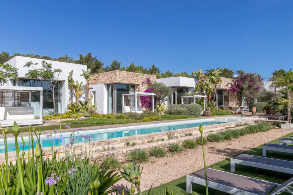 Exterior view of luxury villa Can Paloma featuring modern white architecture, a narrow lap pool, and lush Mediterranean landscaping under a clear blue sky.