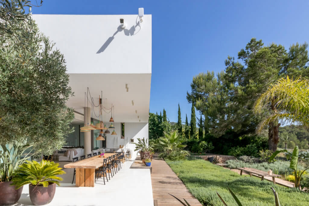Luxury villa Can Paloma featuring a modern outdoor dining area with a long wooden table, eclectic pendant lights, and a lush green Mediterranean garden.