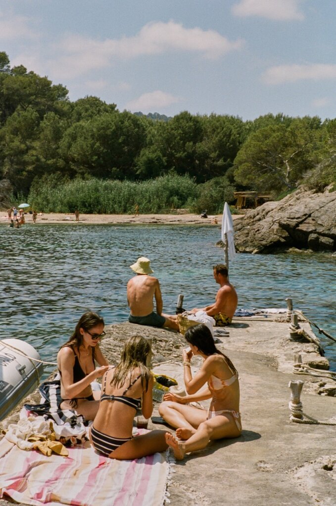 Visitors relax on a rocky shore beside a sheltered swimming cove in Ibiza.