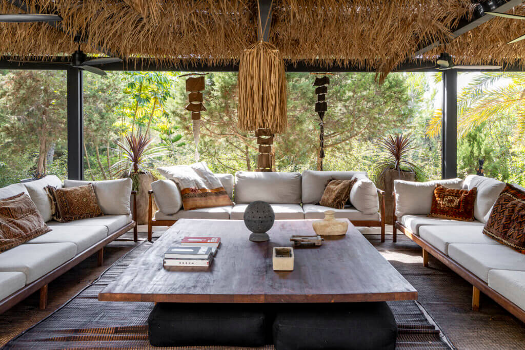 A shaded outdoor lounge at luxury Villa Anaïs featuring grey sofas, ethnic cushions, and a large wooden coffee table under a thatched roof.