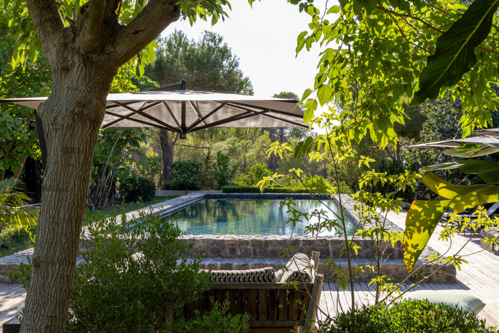 A natural stone-walled swimming pool at luxury Villa Anaïs, framed by lush green trees and a modern poolside umbrella.
