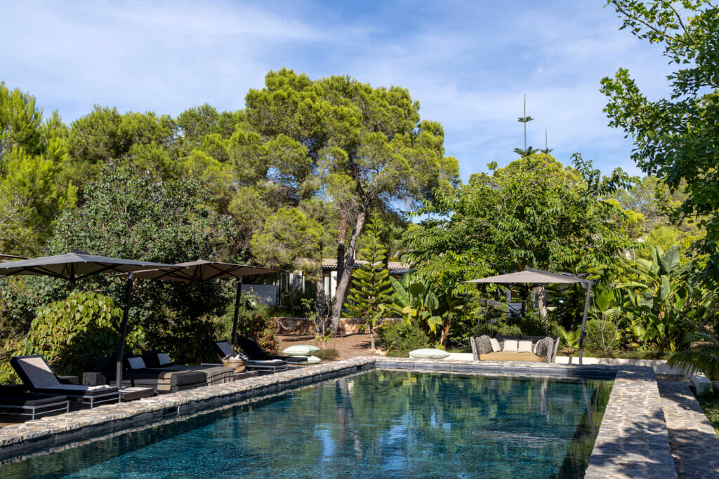 A large stone-edged swimming pool at luxury Villa Anaïs with black sun loungers, grey umbrellas, and a lush background of Mediterranean pine trees.