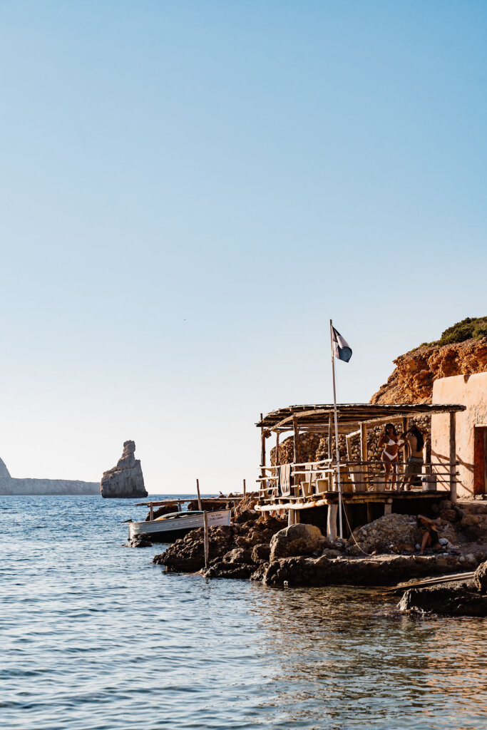 Rocky shore and calm blue water at Benirrás Beach, Ibiza, with a seaside beach bar beside the water.