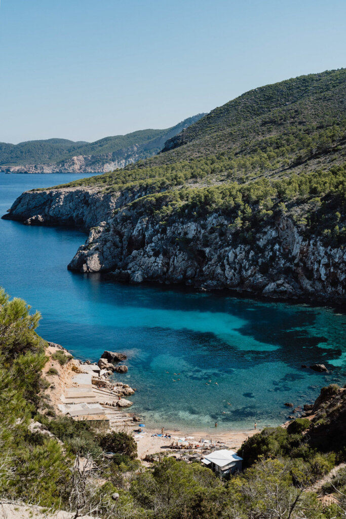 Turquoise water and a small sandy beach below the pine-covered cliffs of Cala d’en Serra, Ibiza.