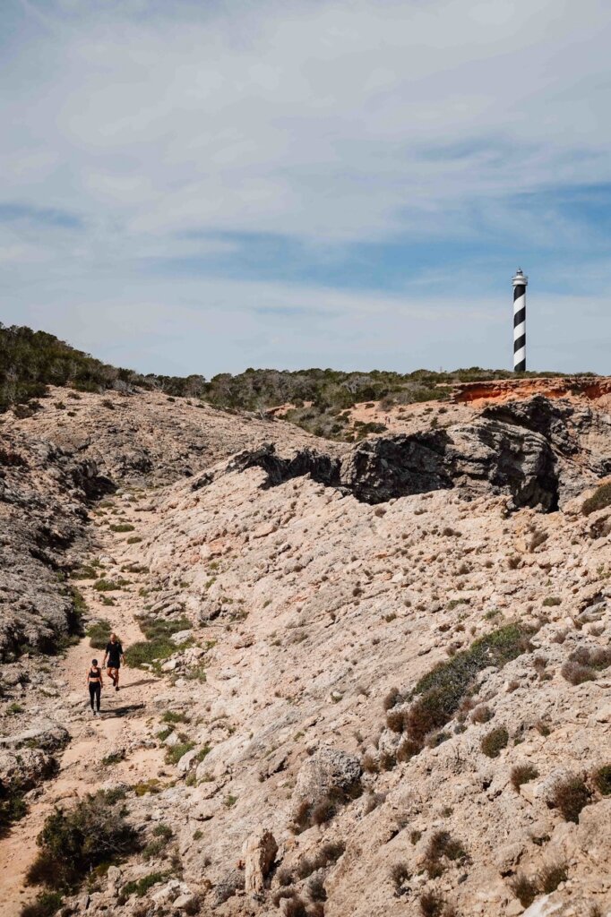 Two hikers follow a rocky trail toward the black-and-white striped Moscarter Lighthouse in Ibiza.