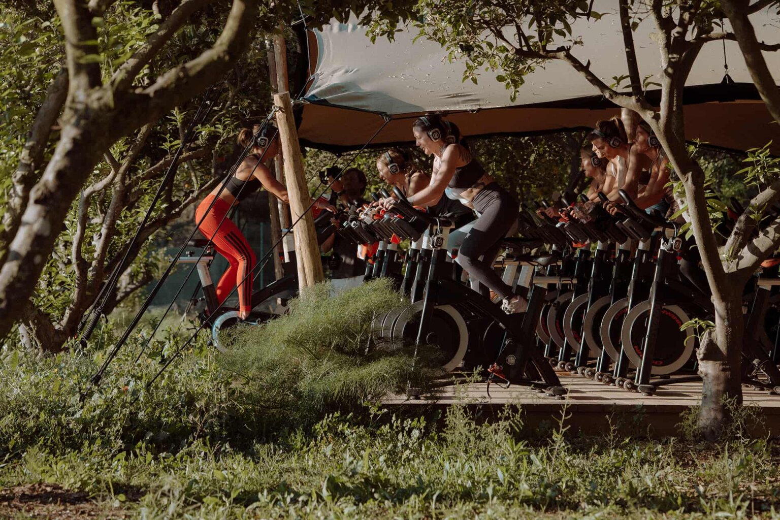Group cycling class on stationary bikes beneath a shaded outdoor canopy at Oranja.